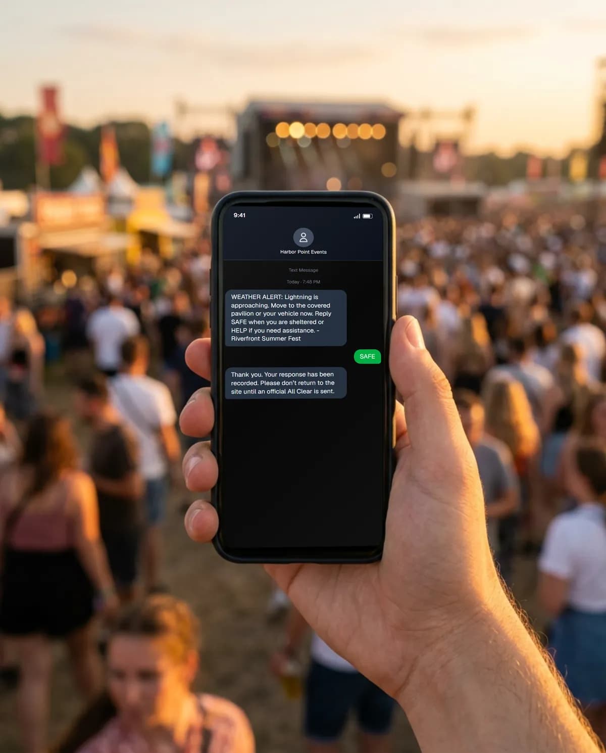 A festival-goer holds up a phone showing a lightning alert from the event, with the crowd and stage behind.